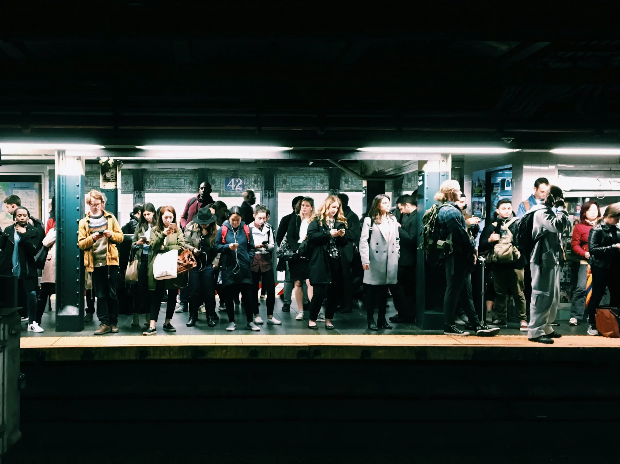 Commuters waiting on a subway platform, some engaged with their phones, during rush hour